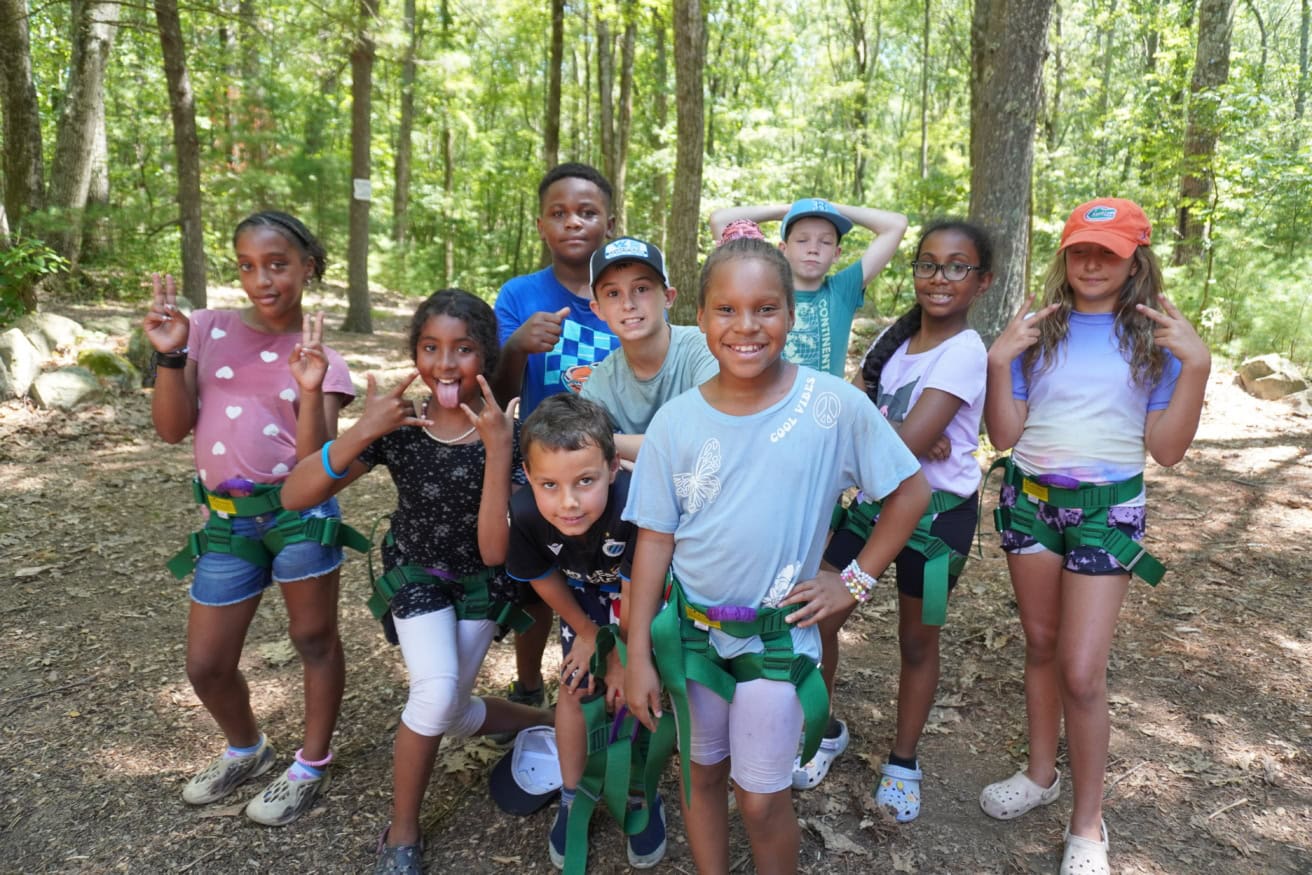 A group of nine children wearing harnesses pose for a photo in a wooded area on a sunny day.