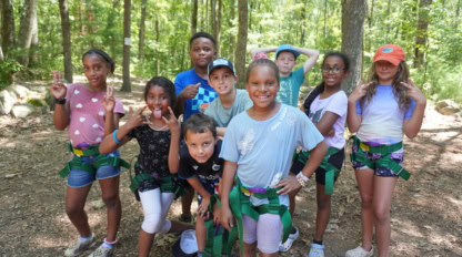 A group of nine children wearing harnesses pose for a photo in a wooded area on a sunny day.