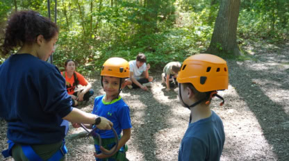 An adult fastens harnesses on two children wearing helmets while other children sit on the ground in a wooded outdoor area.