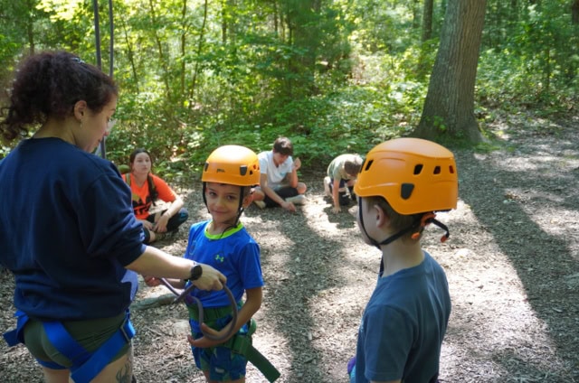 An adult fastens harnesses on two children wearing helmets while other children sit on the ground in a wooded outdoor area.