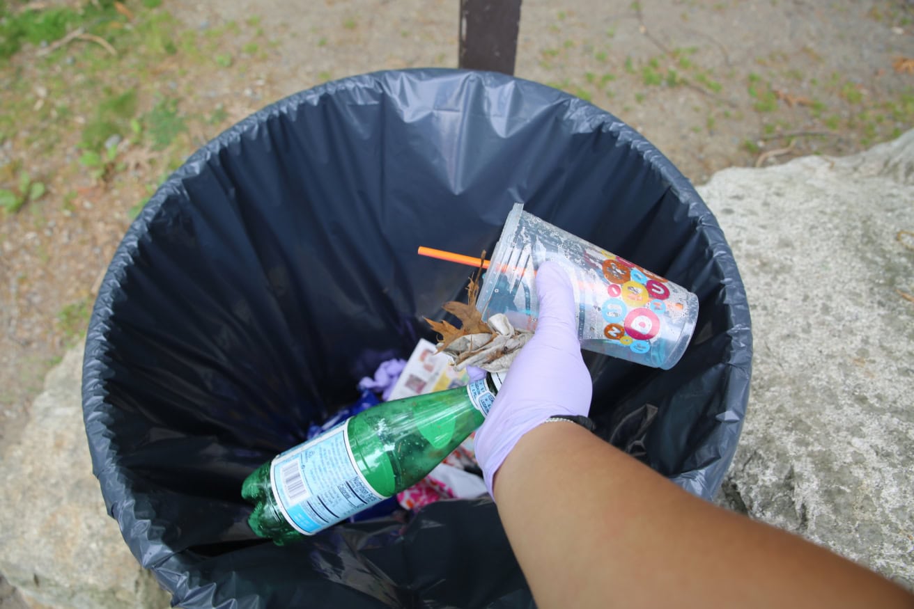 A person wearing a purple glove throws a plastic cup and bottle into a black-lined outdoor trash can.