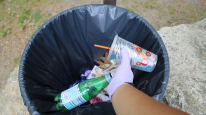 A person wearing a purple glove throws a plastic cup and bottle into a black-lined outdoor trash can.