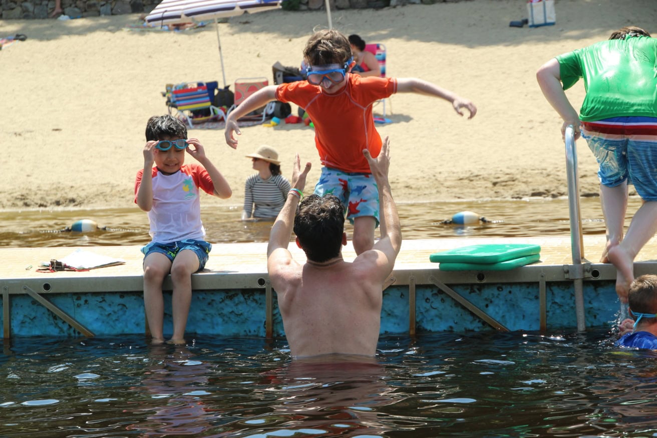 A man in the water reaches up to catch a boy jumping off a dock, while another child sits nearby wearing goggles. People and beach gear are visible in the background.