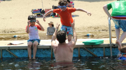 A man in the water reaches up to catch a boy jumping off a dock, while another child sits nearby wearing goggles. People and beach gear are visible in the background.