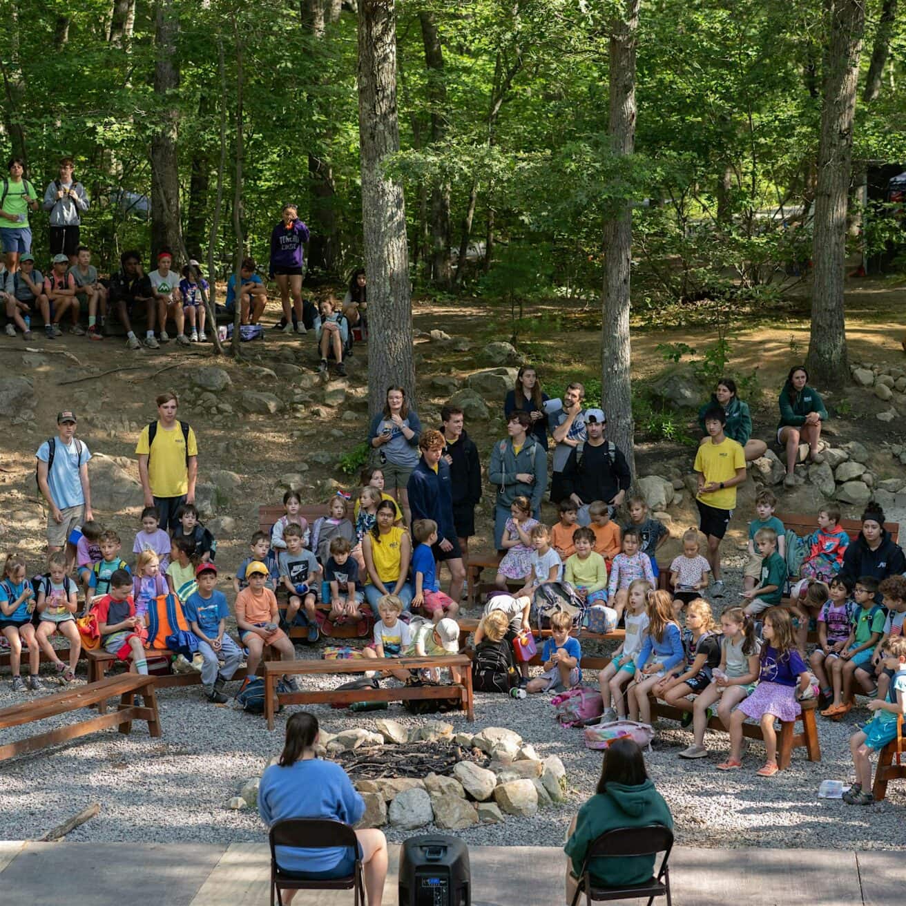 A large group of children and adults sit and stand outdoors in a wooded area, facing two seated individuals near a fire pit in the center.