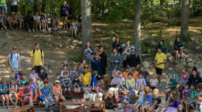 A group of children and adults sit outdoors in a wooded area, gathered around a stone fire pit, with some people seated on benches and others standing.