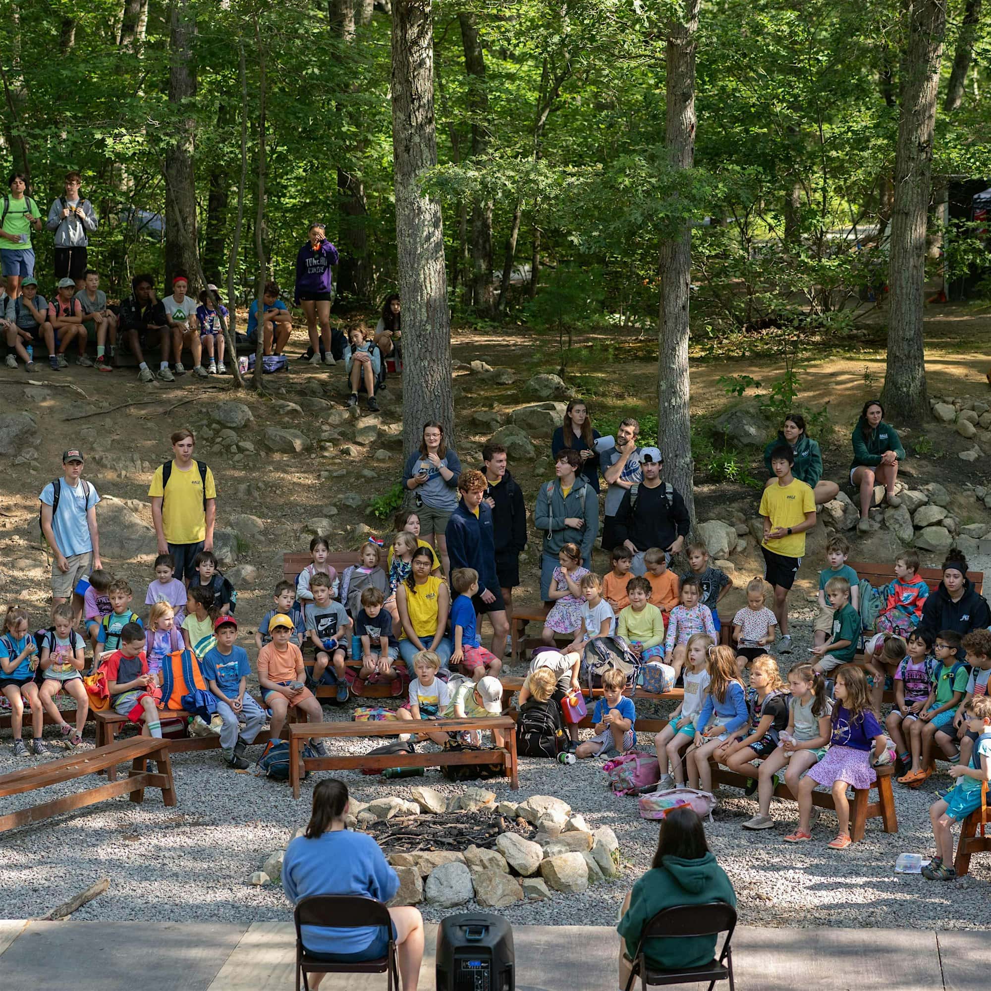 A group of children and adults sit outdoors in a wooded area, gathered around a stone fire pit, with some people seated on benches and others standing.