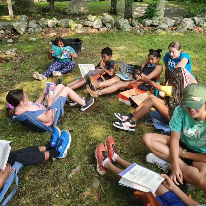 A group of children sit in a grassy clearing reading books, surrounded by trees, with a log cabin in the background.