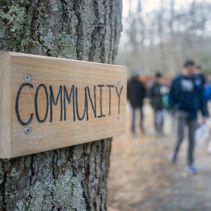 A wooden sign reading "Community" is attached to a tree; a group of people is visible walking along a path in the blurred background.