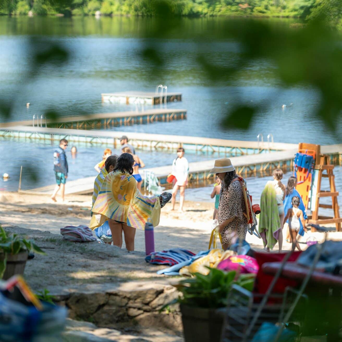 People stand and walk on a sandy lakeshore near wooden docks, some wrapped in towels, with trees and greenery in the background.