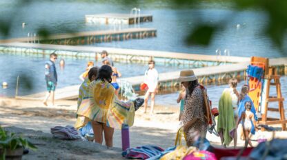 People stand and walk on a sandy lakeshore near wooden docks, some wrapped in towels, with trees and greenery in the background.