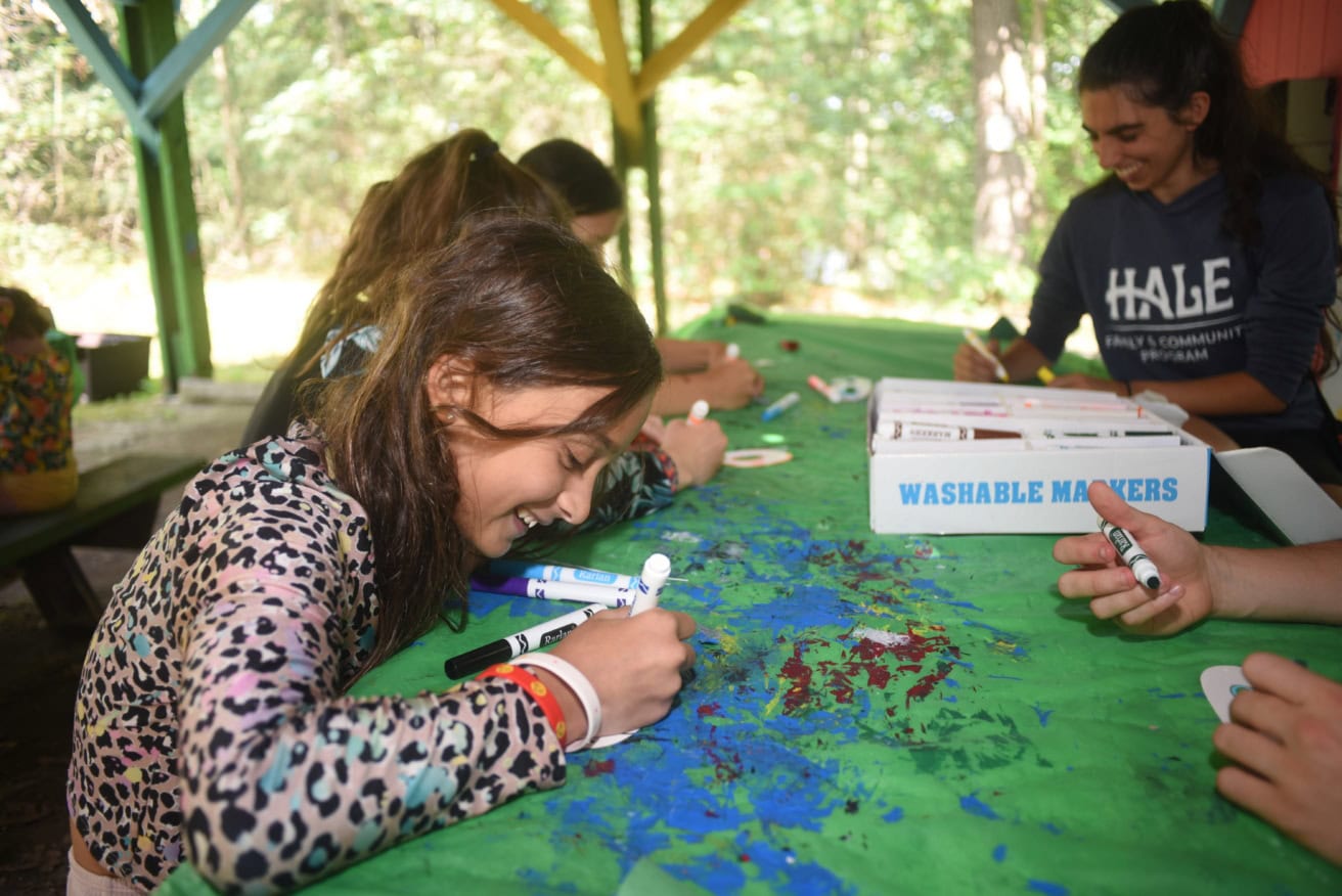 Children sit around a table outdoors, drawing on paper with washable markers. A girl in the foreground smiles as she colors, while others work on their projects in the background.