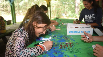 Children sit around a table outdoors, drawing on paper with washable markers. A girl in the foreground smiles as she colors, while others work on their projects in the background.