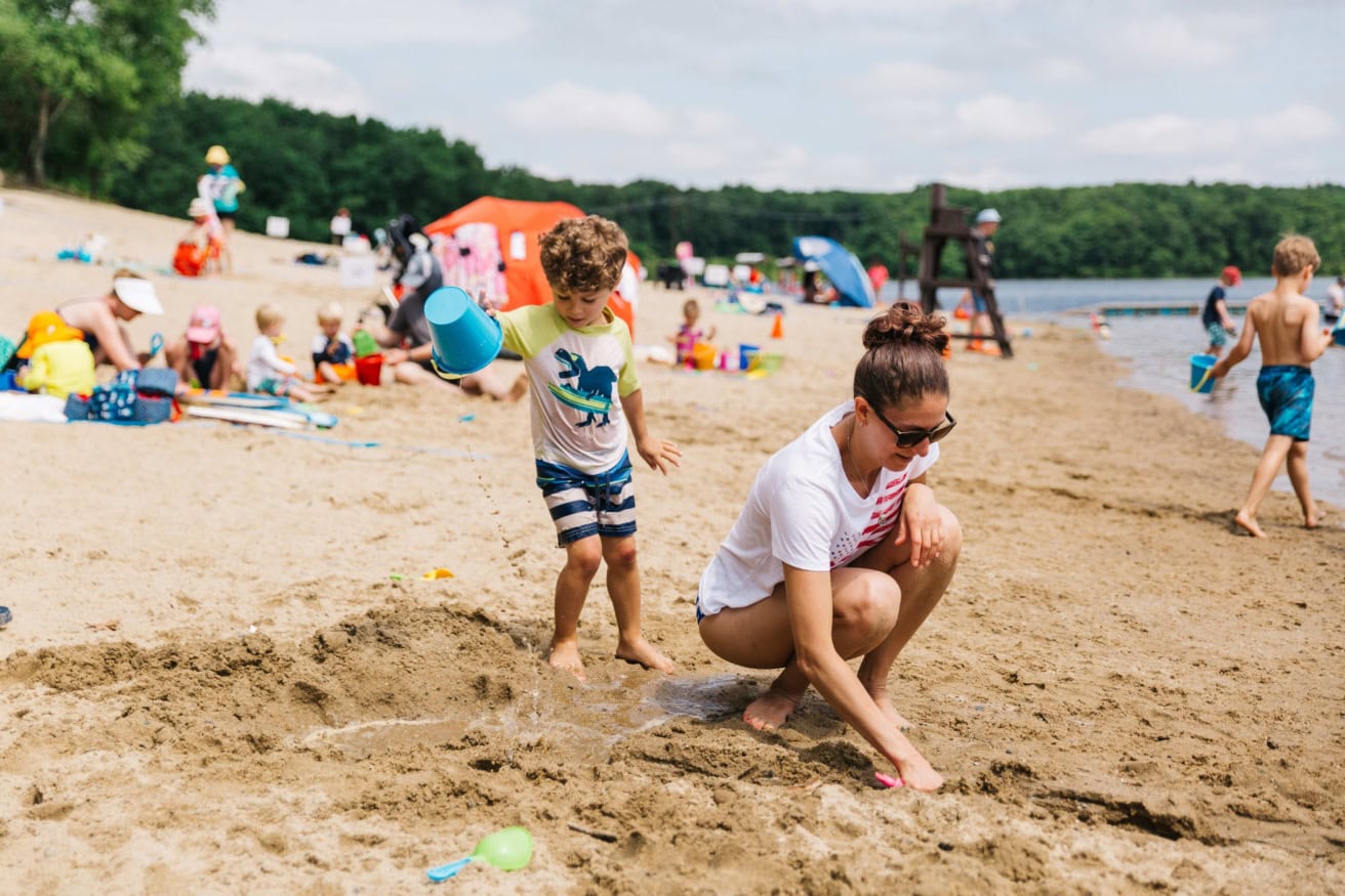 A woman and a young boy play in the sand at a busy lakeside beach surrounded by other people enjoying the water and sun.