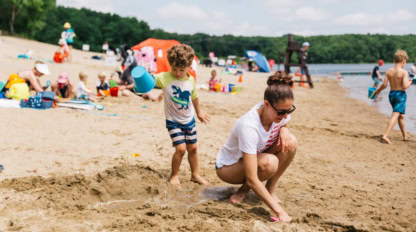 A woman and a young boy play in the sand at a busy lakeside beach surrounded by other people enjoying the water and sun.