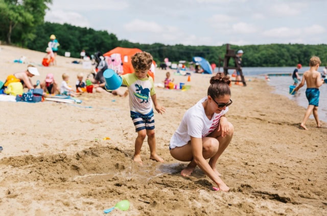 A woman and a young boy play in the sand at a busy lakeside beach surrounded by other people enjoying the water and sun.