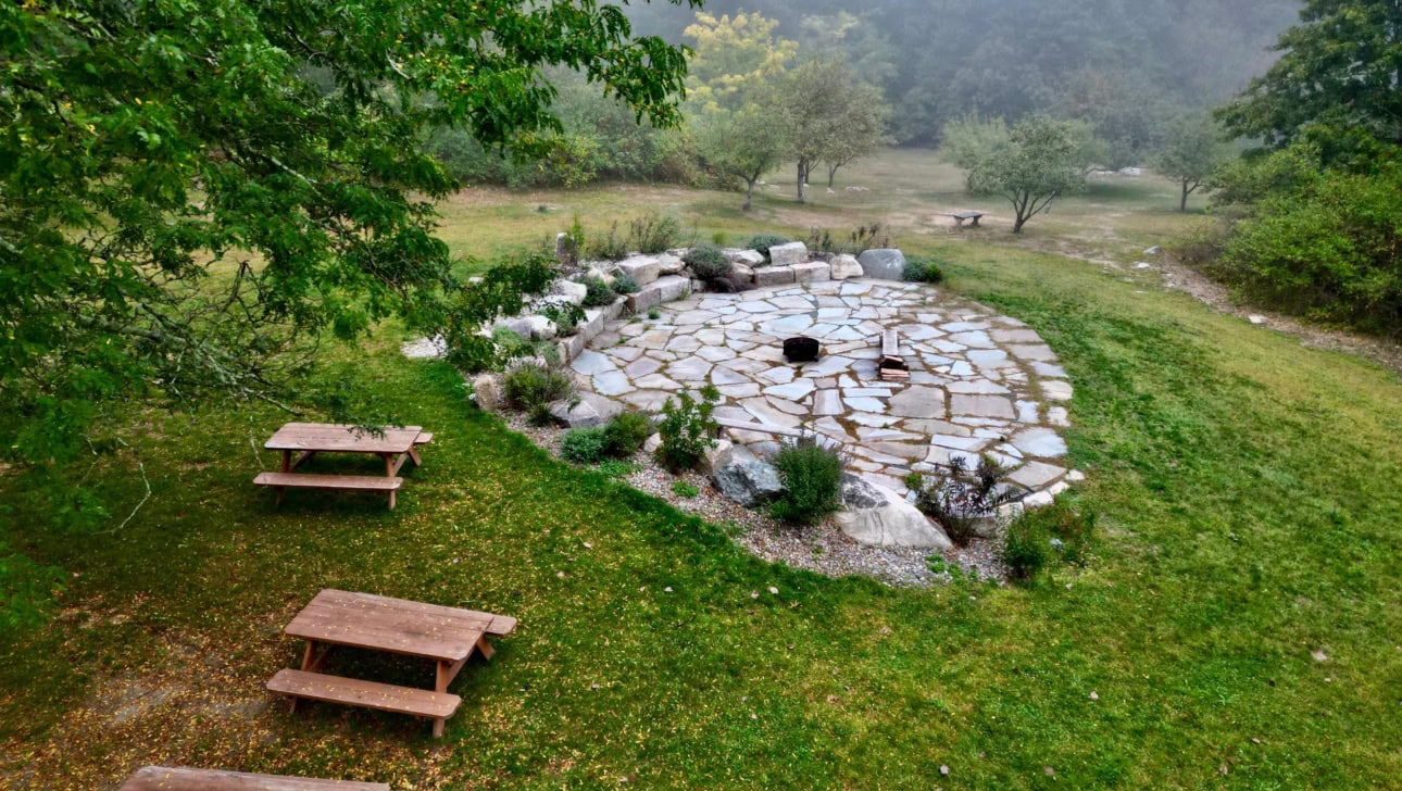 An outdoor stone fire pit with a circle of seating, surrounded by grass, picnic tables, and trees on a foggy day.