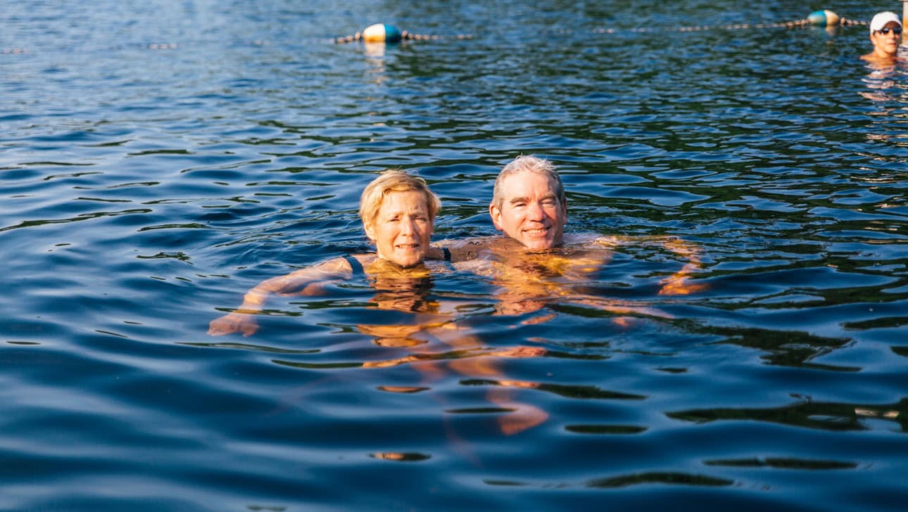 Two older adults are swimming together in a lake, smiling at the camera. Another person is visible in the background, also in the water.
