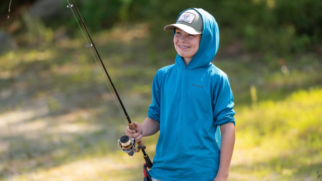 A smiling child wearing a blue hoodie and cap holds a fishing rod outdoors on a sunny day.