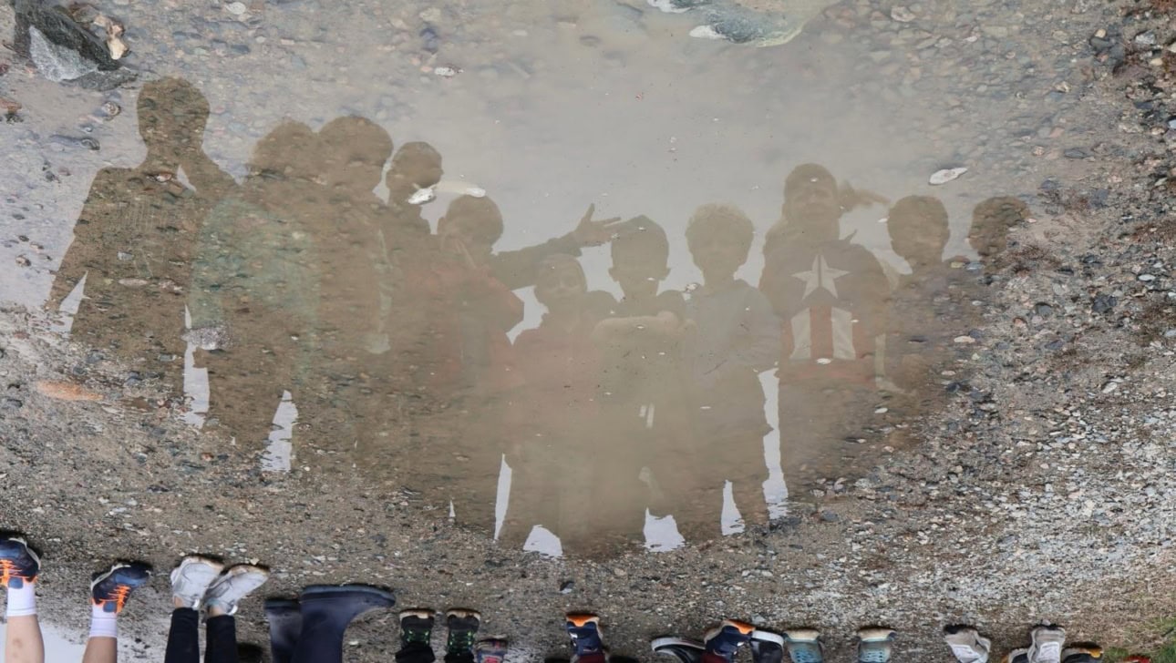 A group of children's reflections are visible in a puddle, while their feet in various shoes and boots are seen at the bottom of the image.