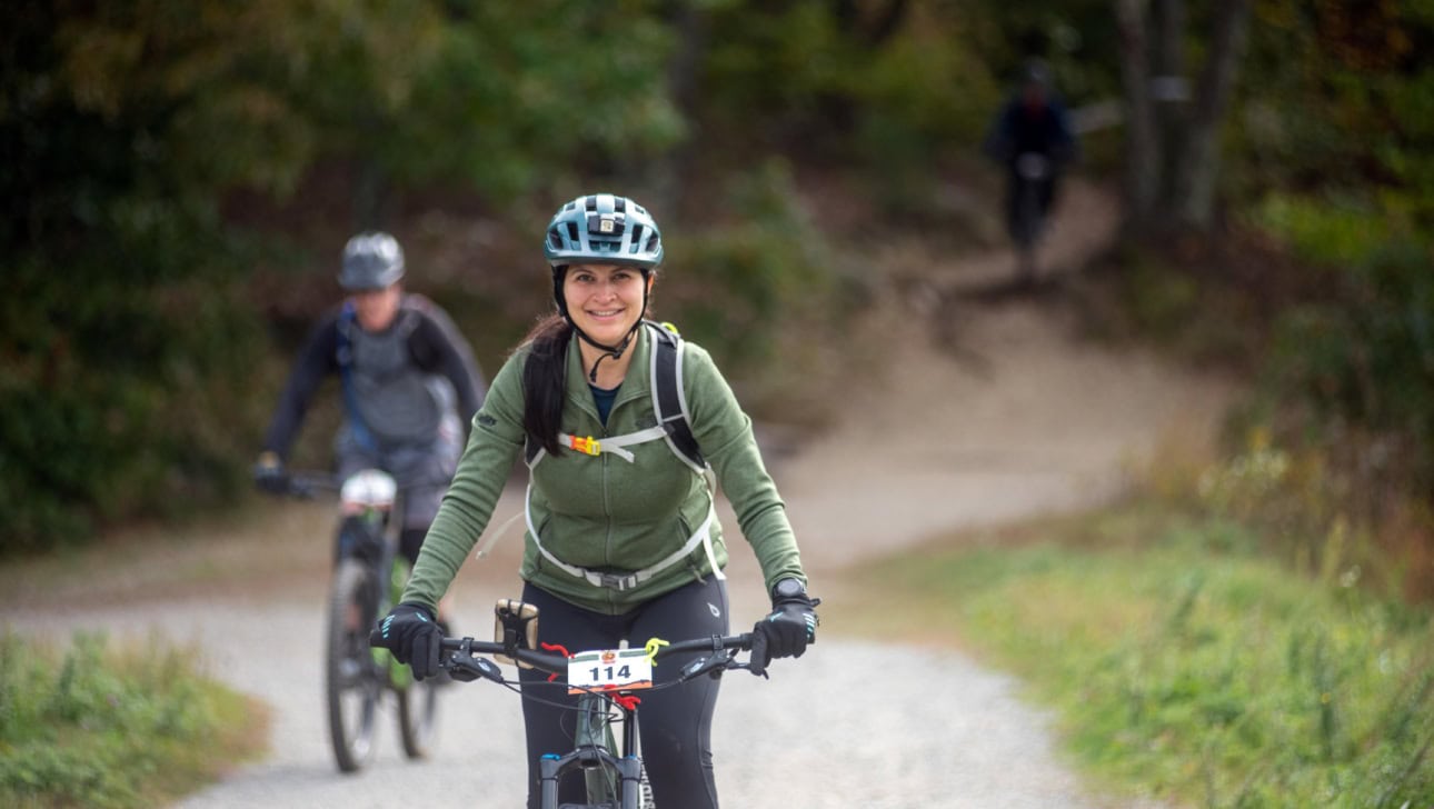 A woman wearing a green jacket and helmet rides a mountain bike on a gravel trail, with another cyclist following behind in a wooded area.