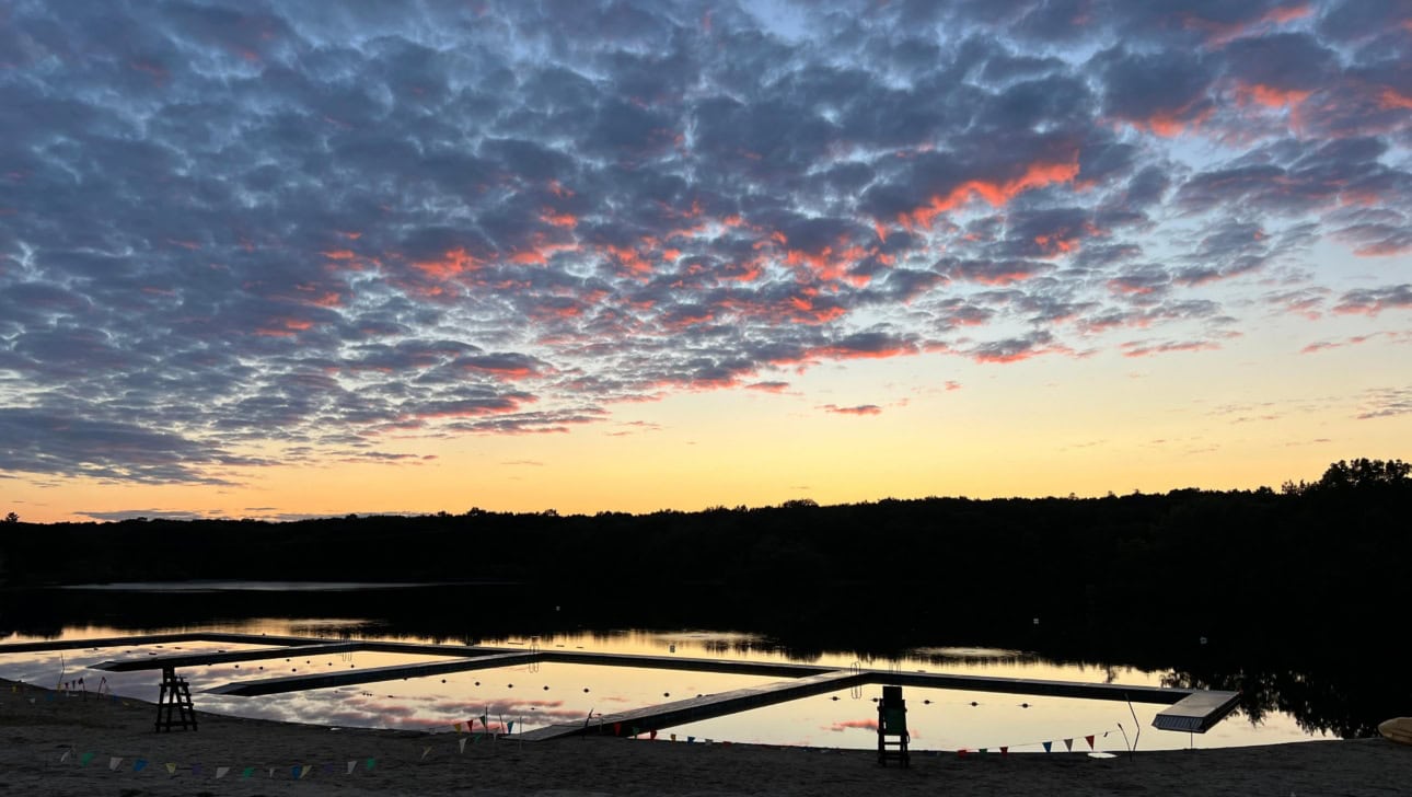 A calm lake at sunset with a sandy shore, lifeguard chairs, floating docks, and a sky filled with clouds tinged with orange.