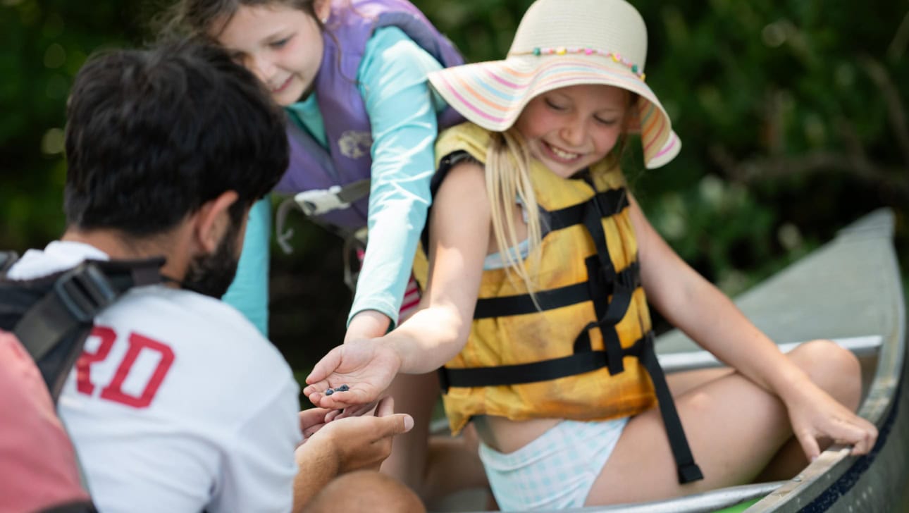 An adult hands a small object to a smiling girl in a life jacket and sun hat while another girl watches, all seated in a canoe outdoors.