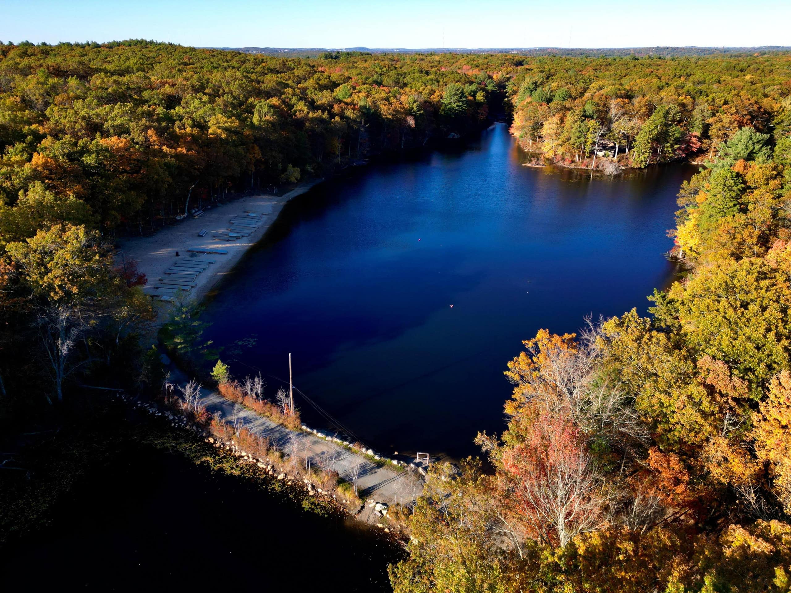 Aerial view of a small lake surrounded by dense autumn foliage, with a sandy beach and boats lined up on the left shoreline.