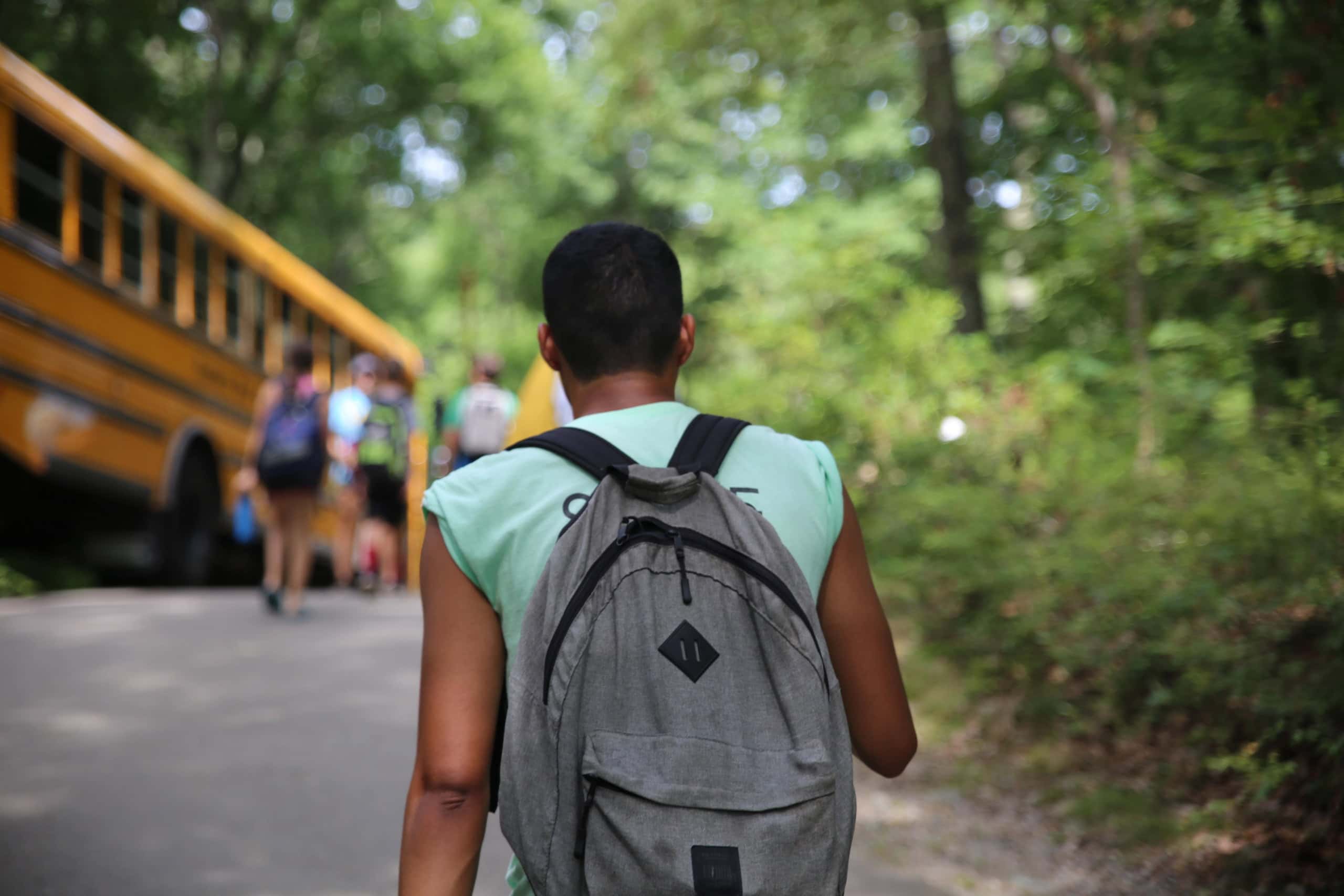 A person with a gray backpack walks on a path toward a group of people and a yellow school bus in a wooded area.
