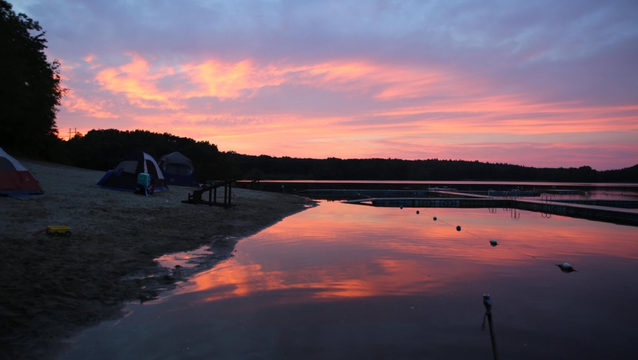 A lakeside campsite at sunset with tents on the sandy shore, docks on the water, and colorful reflections of the sky in the calm lake.