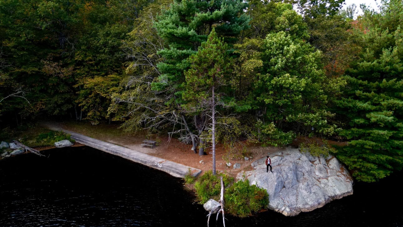 A person stands on a rocky ledge by a lake, surrounded by dense trees, with a picnic table visible nearby.