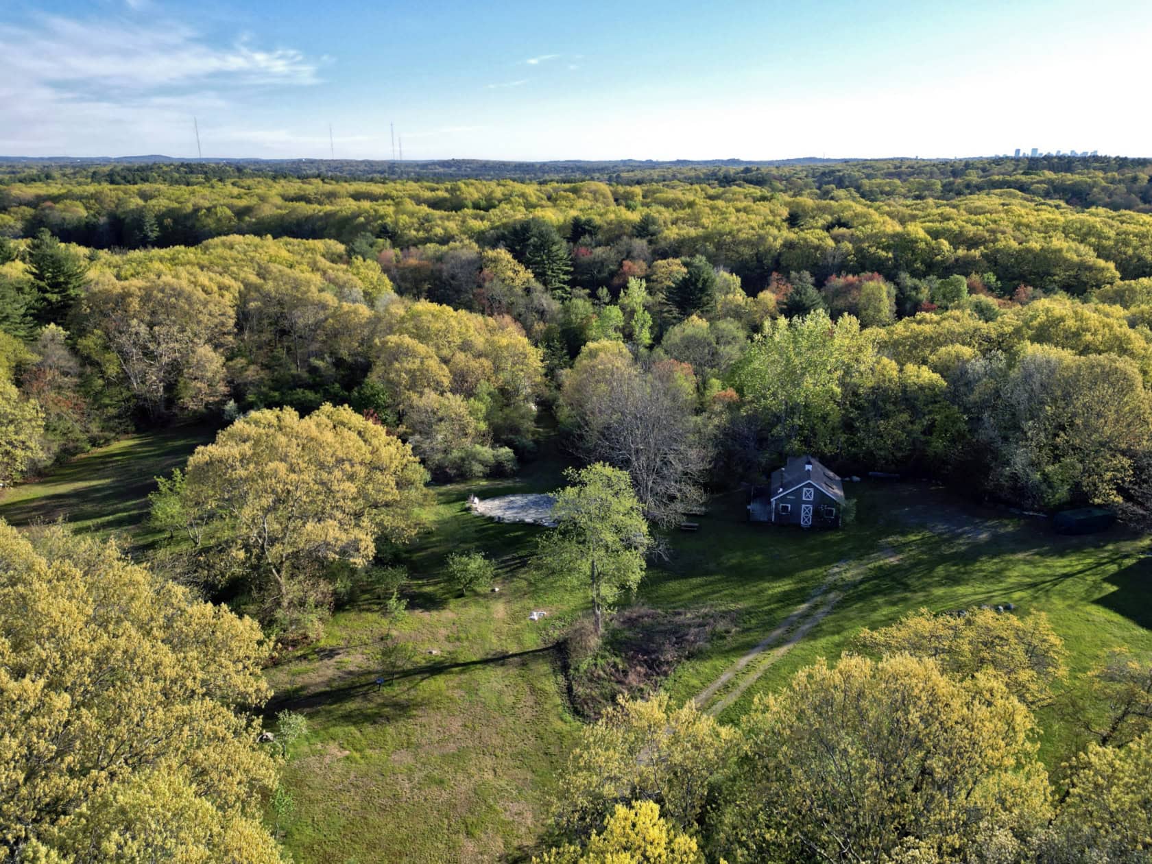 Aerial view of a small house surrounded by dense green forest and open grassy areas under a clear blue sky.