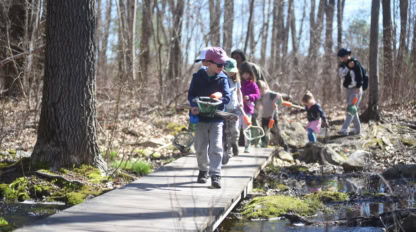 Children and adults walk on a wooden path through a forest carrying nets and buckets on a sunny day, participating in conservation activities that help protect the natural environment.