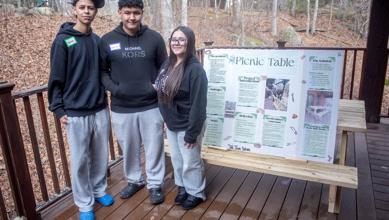 Three people stand on a wooden deck next to a project display board titled “Picnic Table,” which features text, photos, and diagrams. Trees with fallen leaves are visible in the background.