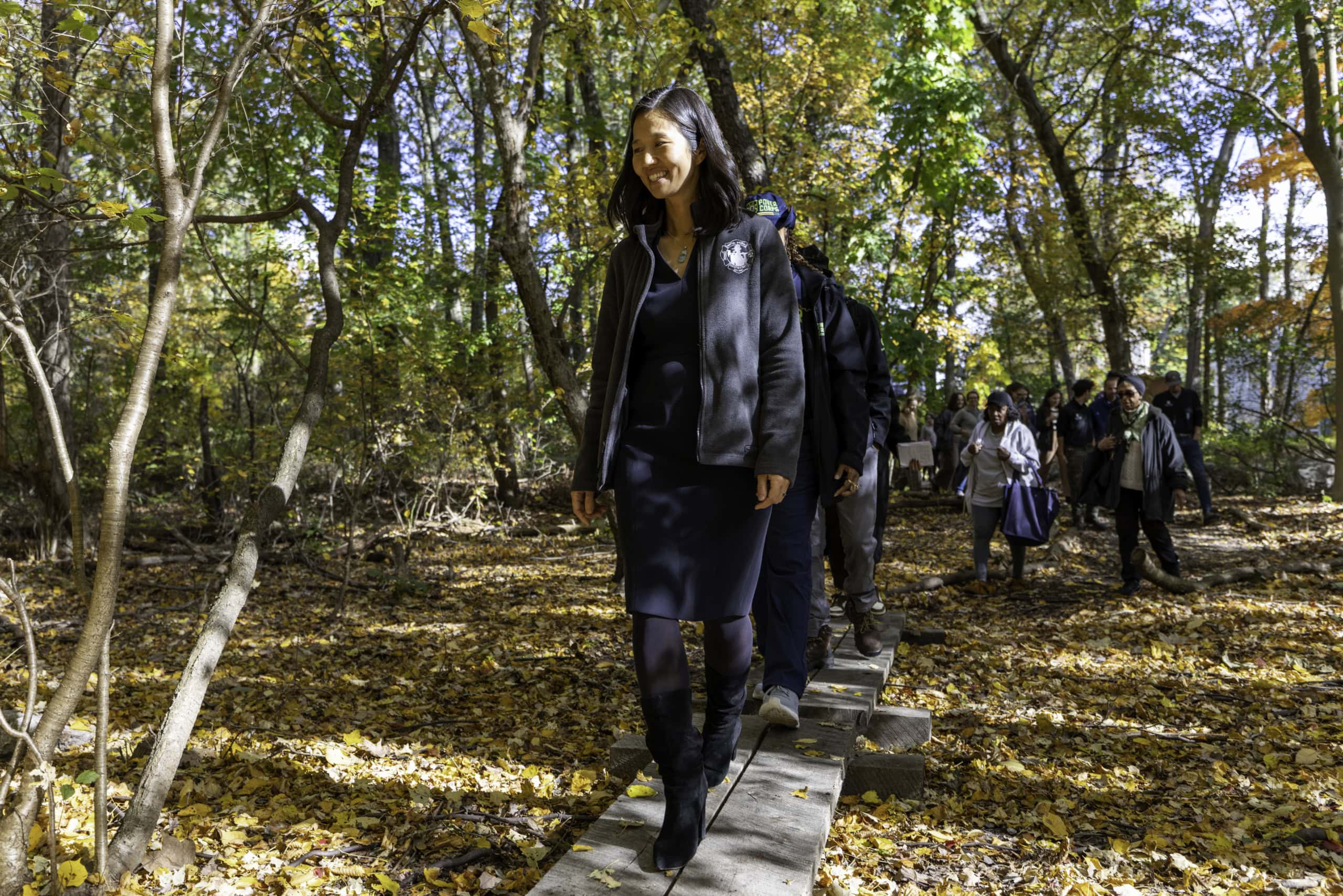 A group of people walk on a narrow path through a sunlit, wooded area covered with fallen autumn leaves, appreciating the beauty of nature and the importance of conservation.