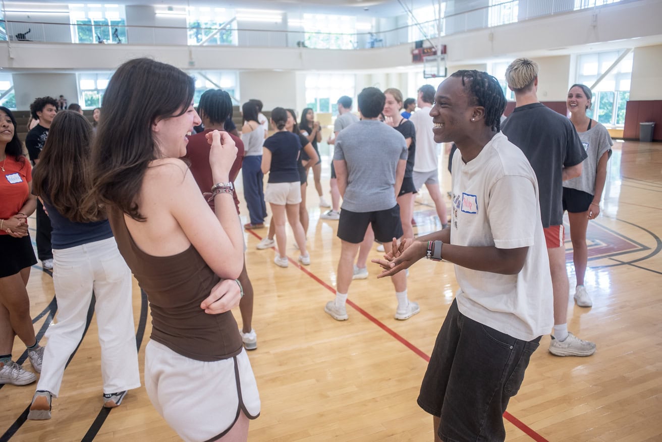 students in a gym do teambuilding activities. Two young students in the front are laughing together.