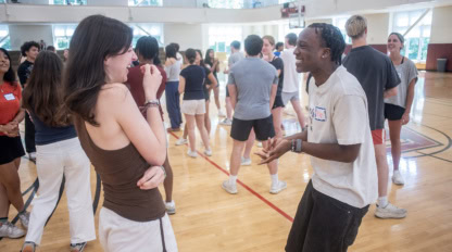 students in a gym do teambuilding activities. Two young students in the front are laughing together.