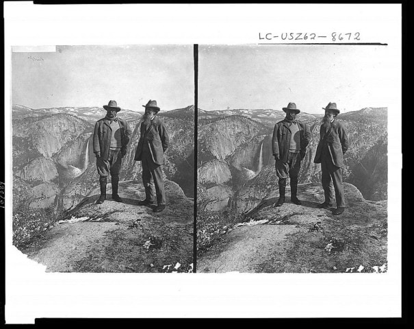 Two men in hats and outdoor clothing stand on a rocky cliff overlooking a mountainous landscape with distant waterfalls.